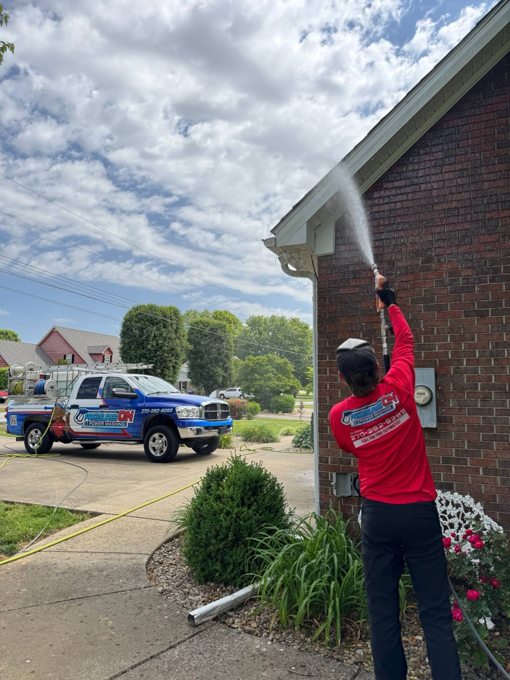 A Pressures ON crew member washing the eaves of a brick house, with the branded truck in the driveway